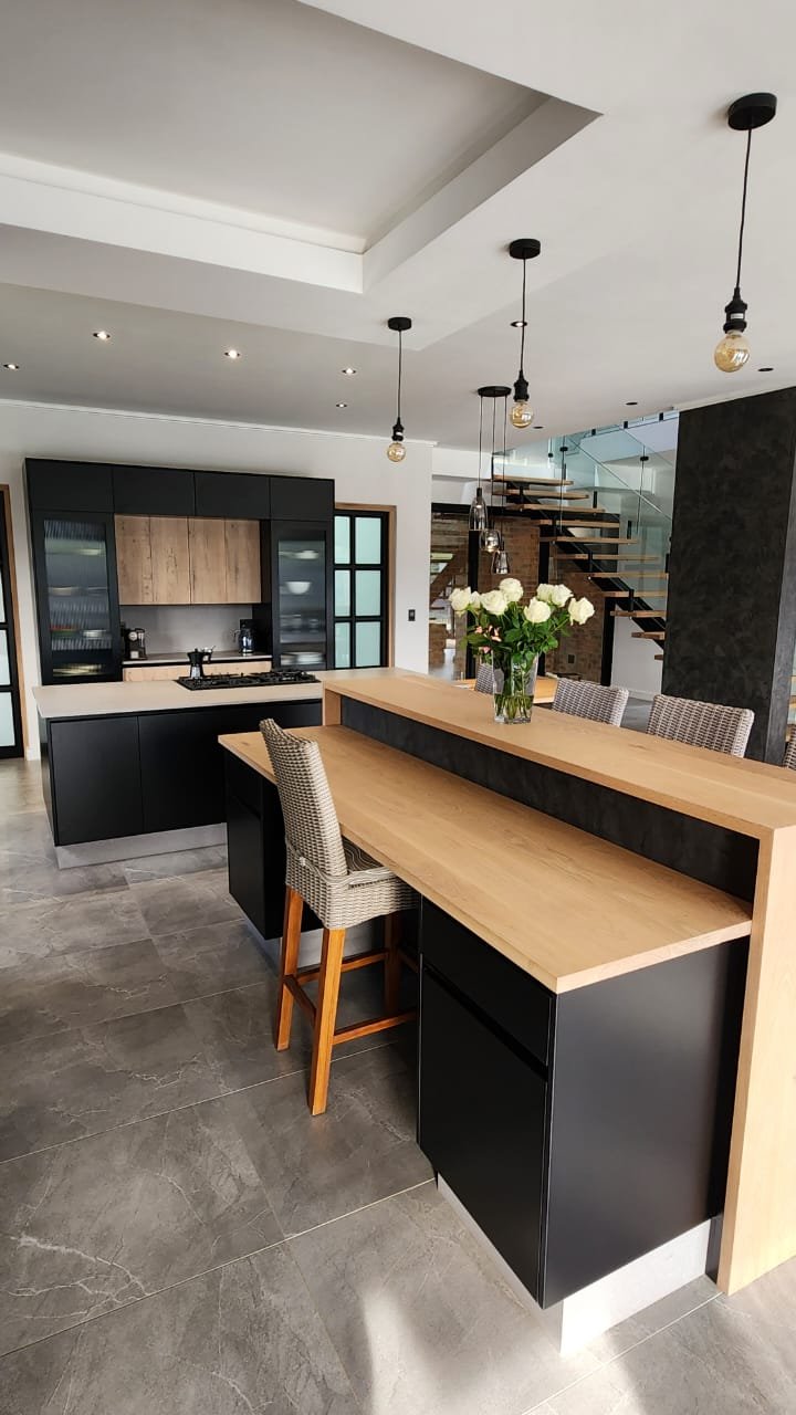 Open-plan kitchen with black cabinetry, a floating oak breakfast bar and a glass-balustrade staircase beyond