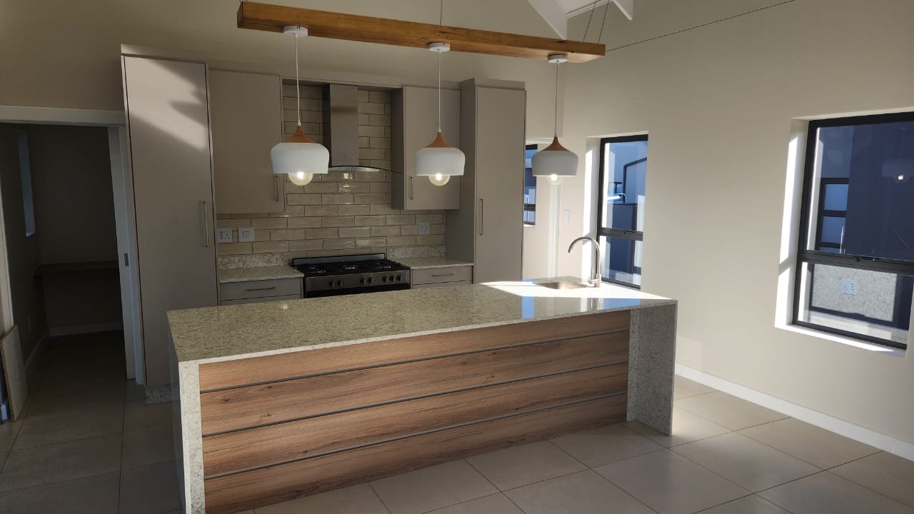 A bright open-plan kitchen with stone-topped wood island, exposed beam pendant and brick splashback