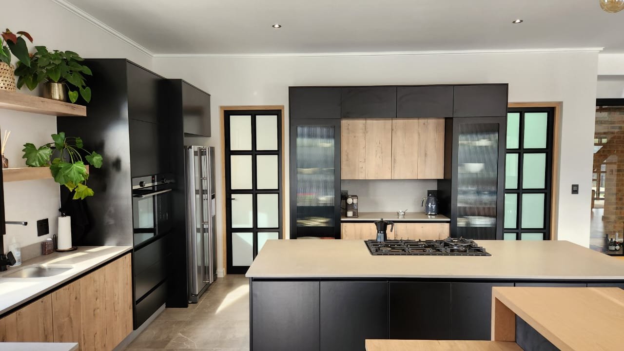 A wide black-and-oak open-plan kitchen with frosted steel-frame doors, stone island and paired pantry